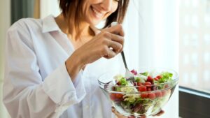 A woman in a white shirt smiling while holding a glass bowl of fresh salad with cherry tomatoes, leafy greens, and using a fork to eat, standing near a bright window.
