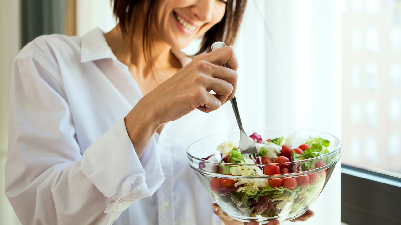 A woman in a white shirt smiling while holding a glass bowl of fresh salad with cherry tomatoes, leafy greens, and using a fork to eat, standing near a bright window.