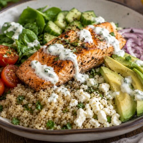 Mediterranean salmon quinoa bowl with roasted salmon, avocado, cherry tomatoes, cucumber, spinach, feta cheese, and creamy herb yogurt sauce in a rustic ceramic bowl