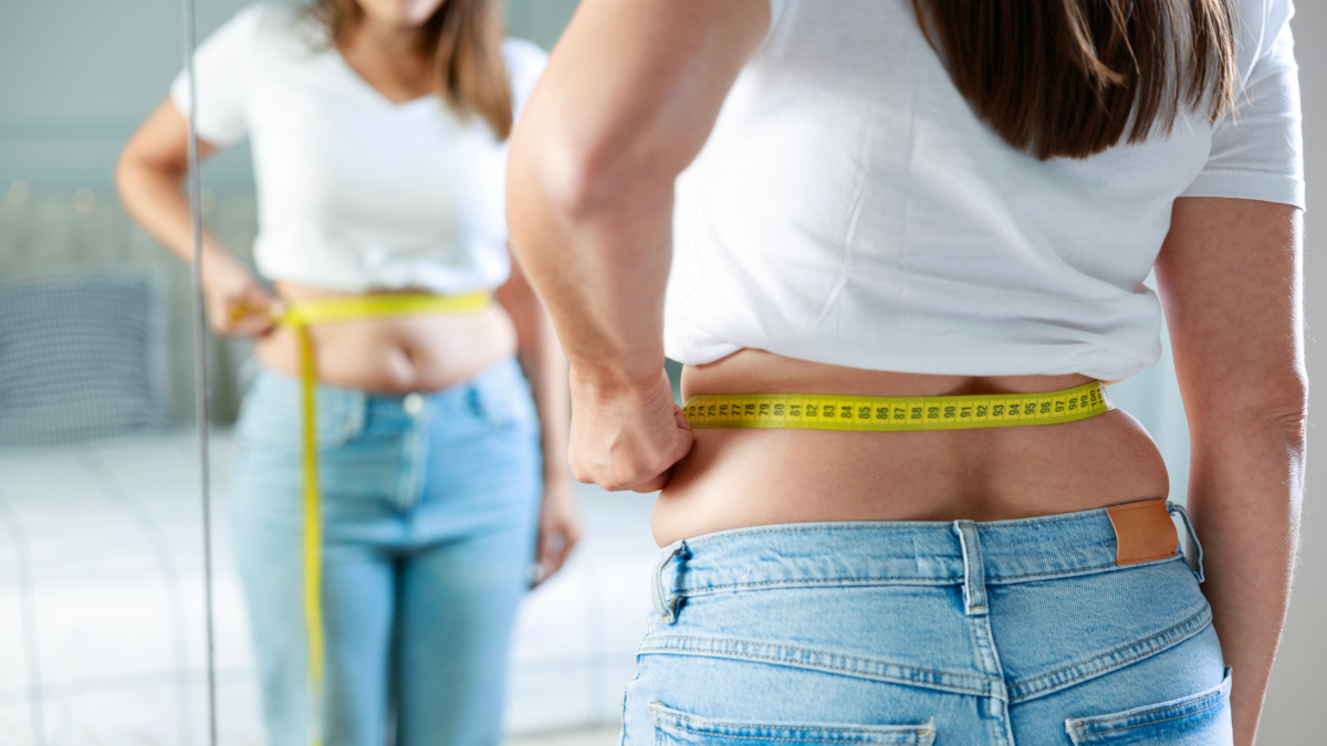 Midlife woman measuring her waist in front of a mirror while reflecting on menopause-related body changes and weight gain.