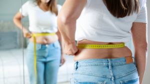 Midlife woman measuring her waist in front of a mirror while reflecting on menopause-related body changes and weight gain.