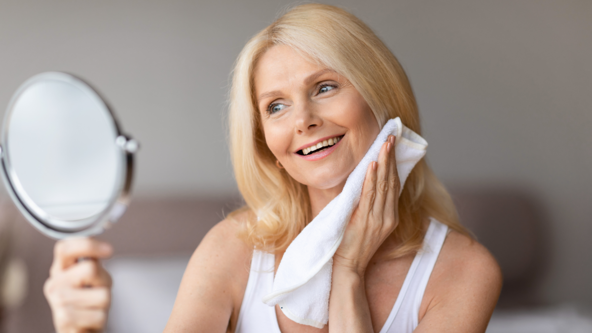 Smiling midlife woman gently touching her face with a towel while looking in a mirror during her skincare routine.