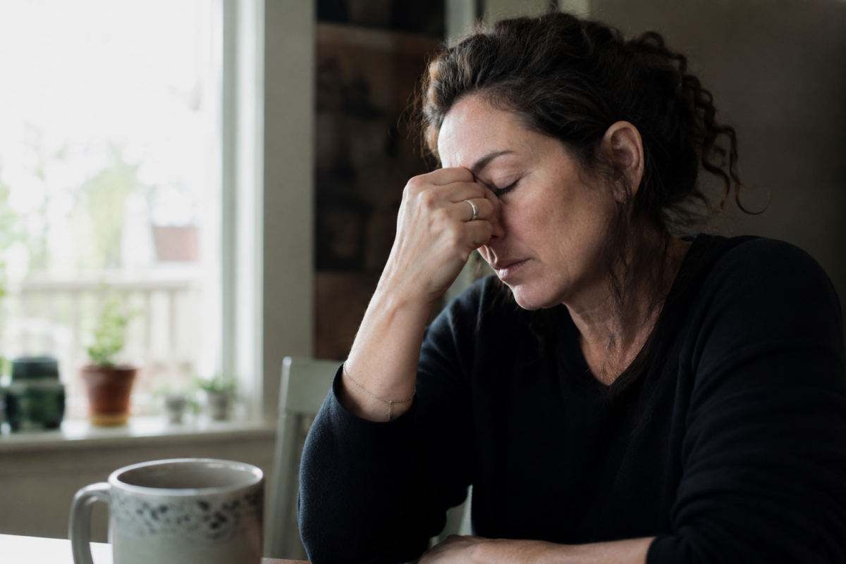 A thoughtful middle-aged woman sits at a kitchen table beside a coffee mug, resting her face in her hand as she reflects quietly in soft morning light.