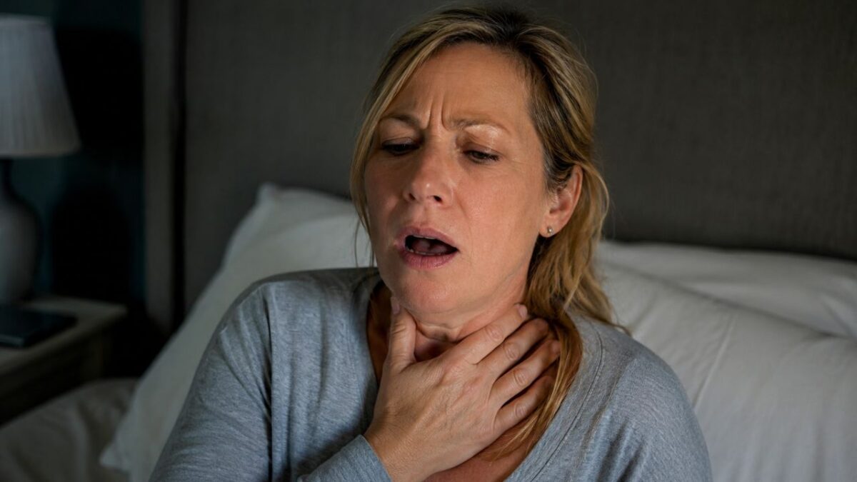 Midlife woman sitting up in bed holding her throat after suddenly choking on her saliva during the night, illustrating a possible perimenopause-related symptom.